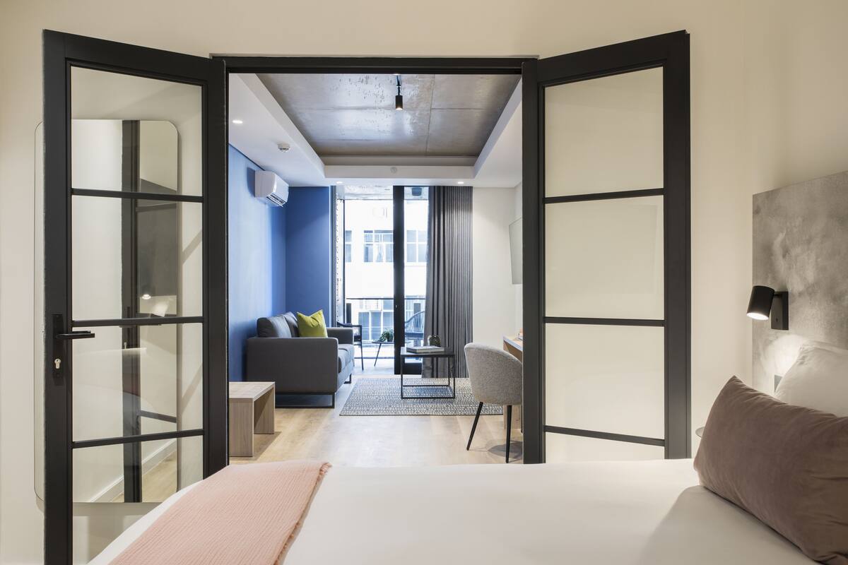 Habitación de hotel moderna con cama, escritorio, silla y sofá. Se ve la ciudad a través de ventanas amplias.