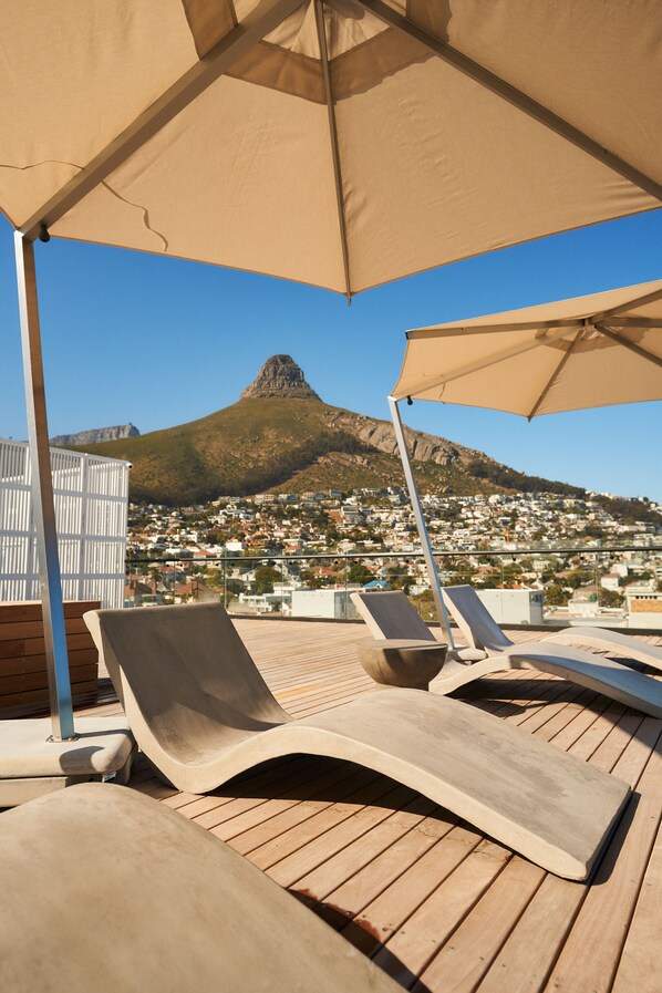 A wooden deck with lounge chairs and a large umbrella, overlooking a town and a mountain.