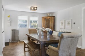 A dining room with a wooden table, chairs, a bench, a radiator, a window, a cabinet, and wall-mounted artwork.