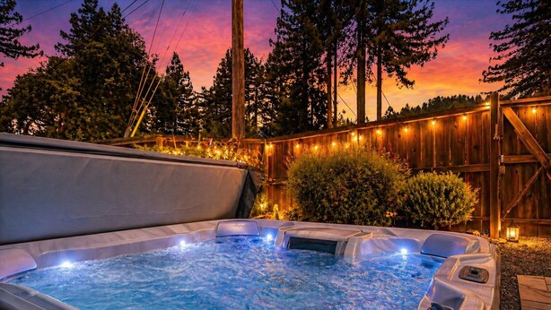 Un bain à remous doté d’une terrasse en bois et illuminé de guirlandes lumineuses, entouré d’arbres et délimité par une clôture en bois.