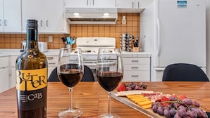 Une bouteille de vin, deux verres de vin rouge et une planche de charcuterie avec du fromage et des fruits, le tout dans une cuisine moderne.
