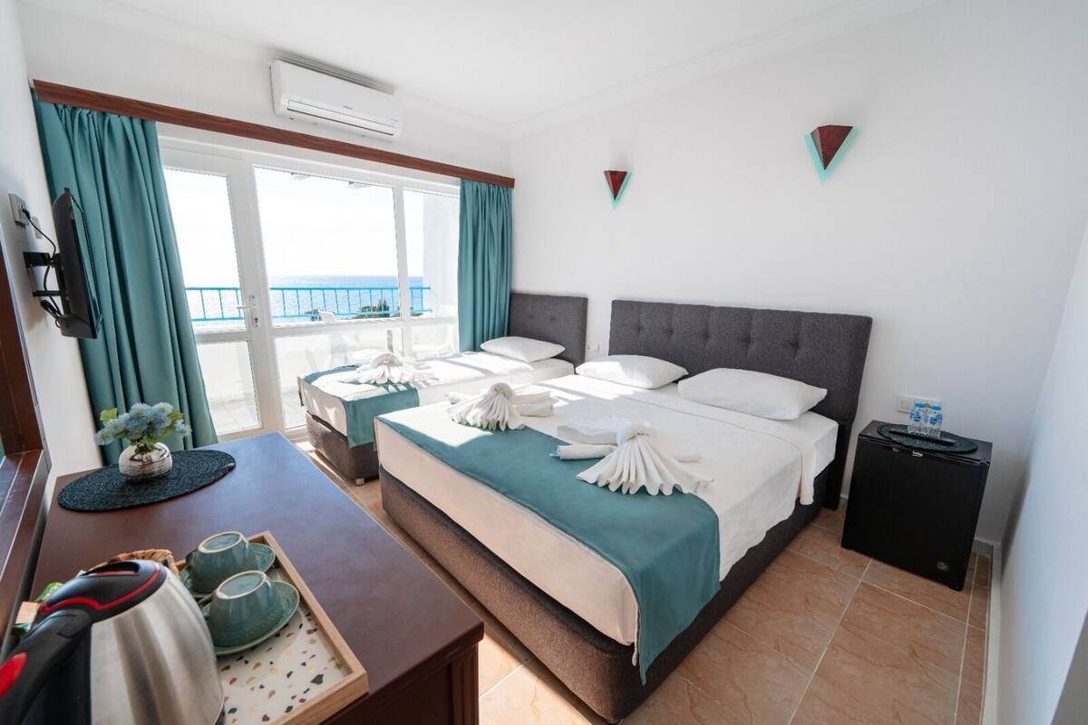 Habitación de hotel con dos camas, un escritorio con hervidor, una mesita con tazas y vista al mar.