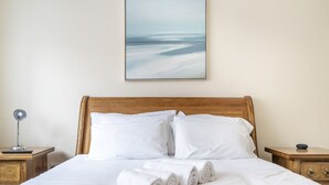 Ein ordentlich bezogenes Bett mit weißen Leinen und aufgerollten Handtüchern, ein hölzerner Kopfteil und ein gerahmtes Bild an der Wand.