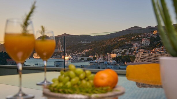 A table with two glasses of orange juice, a plate of green grapes, and an orange, with a view of a city and mountains in the background. - Hedera Estate, Hedera A15 (Dubrovnik)