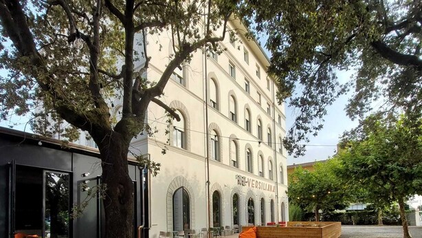 An outdoor dining area with wooden tables and chairs, a large tree, and a building with arched windows in the background. - RE-Versiliana (Pietrasanta)