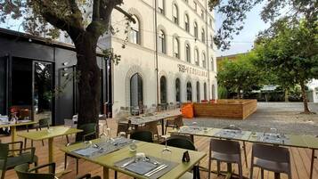 Area pranzo all'aperto con tavoli e sedie in legno, un grande albero e un edificio con finestre ad arco sullo sfondo.