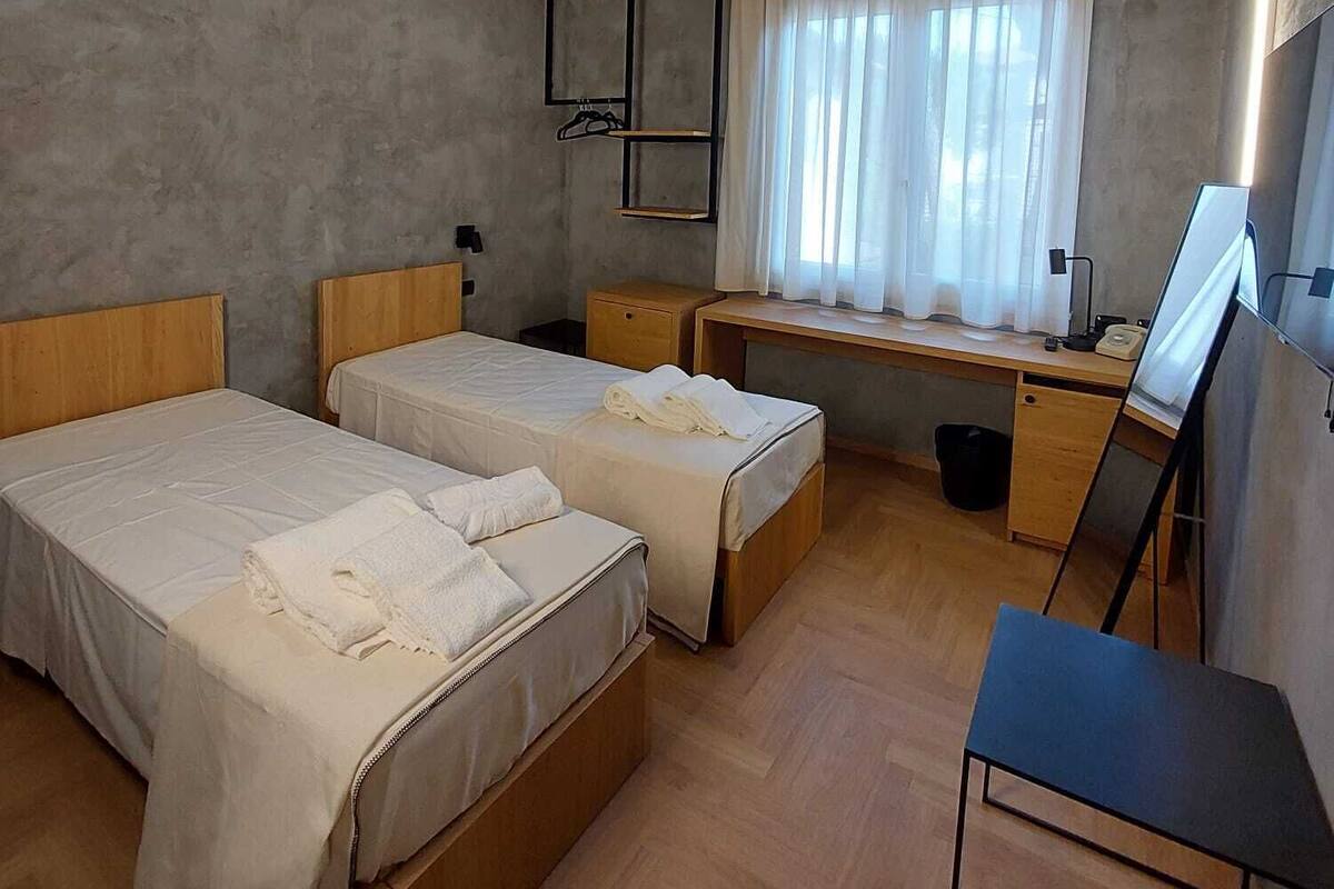 A room with two single beds, a desk with a chair, a large window with curtains, and a mirror on the wall.
