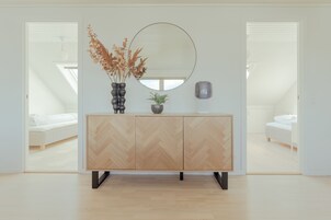 Un dormitorio moderno con un aparador de diseño geométrico tipo espiga, un espejo redondo y un jarrón con flores secas.