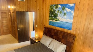 Une pièce aux murs lambrissés, comprenant un lit, une table de chevet avec une lampe, une porte et un tableau représentant une scène de plage.