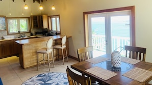 Une cuisine avec des meubles en bois, un coin bar et une table à manger avec des chaises. Une grande porte coulissante offre une vue sur l’océan.