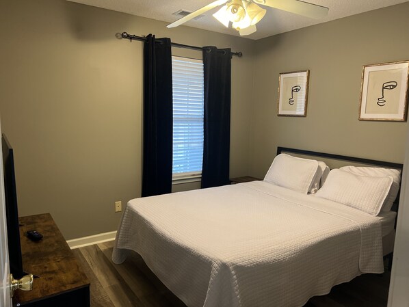 Ein Schlafzimmer mit einem Bett, einem Fenster mit Vorhängen, einem Deckenventilator und zwei gerahmten Bildern an der Wand.