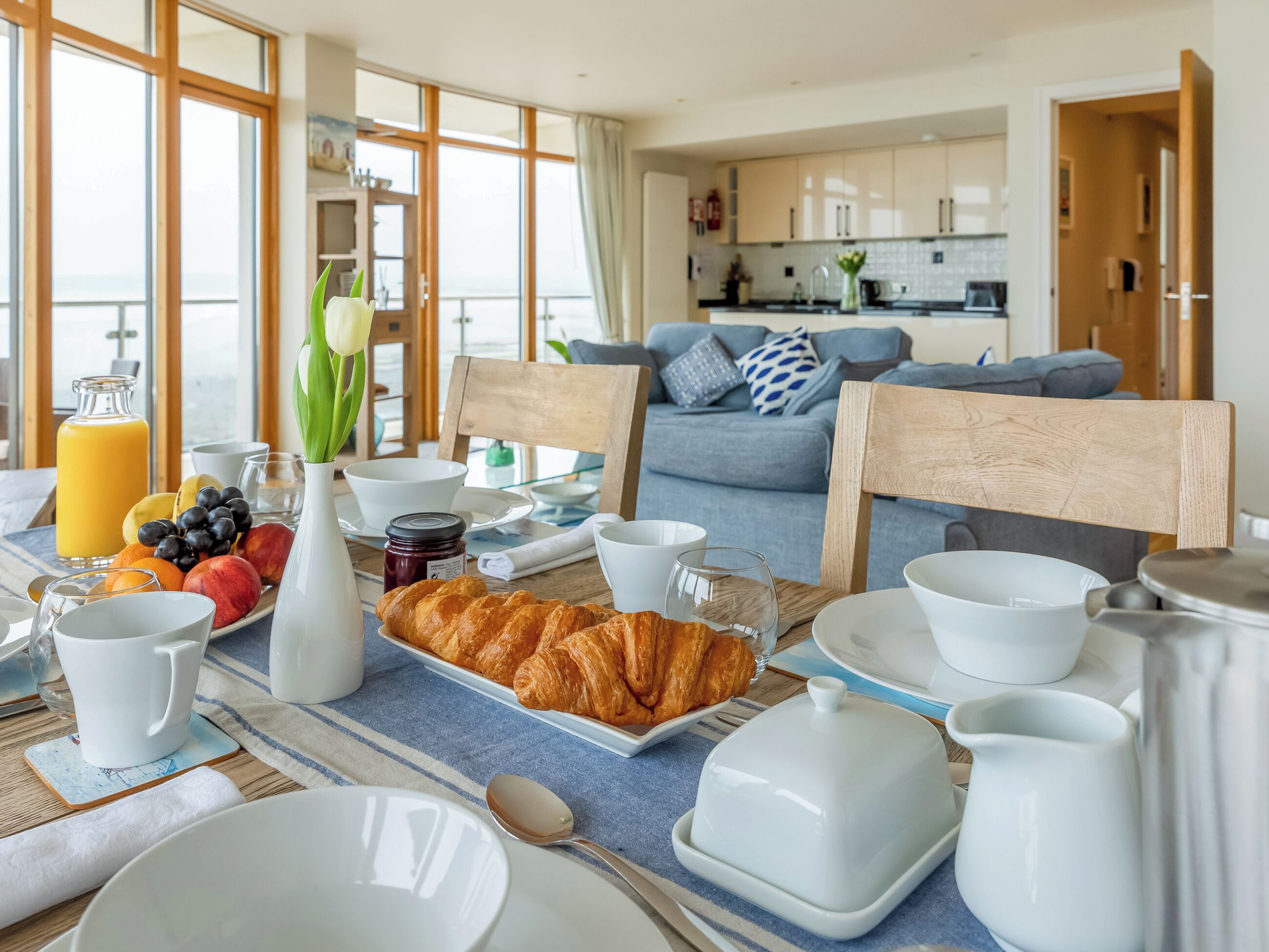 Une table dressée pour le petit-déjeuner, avec des croissants, des fruits et une bouteille de jus d’orange.