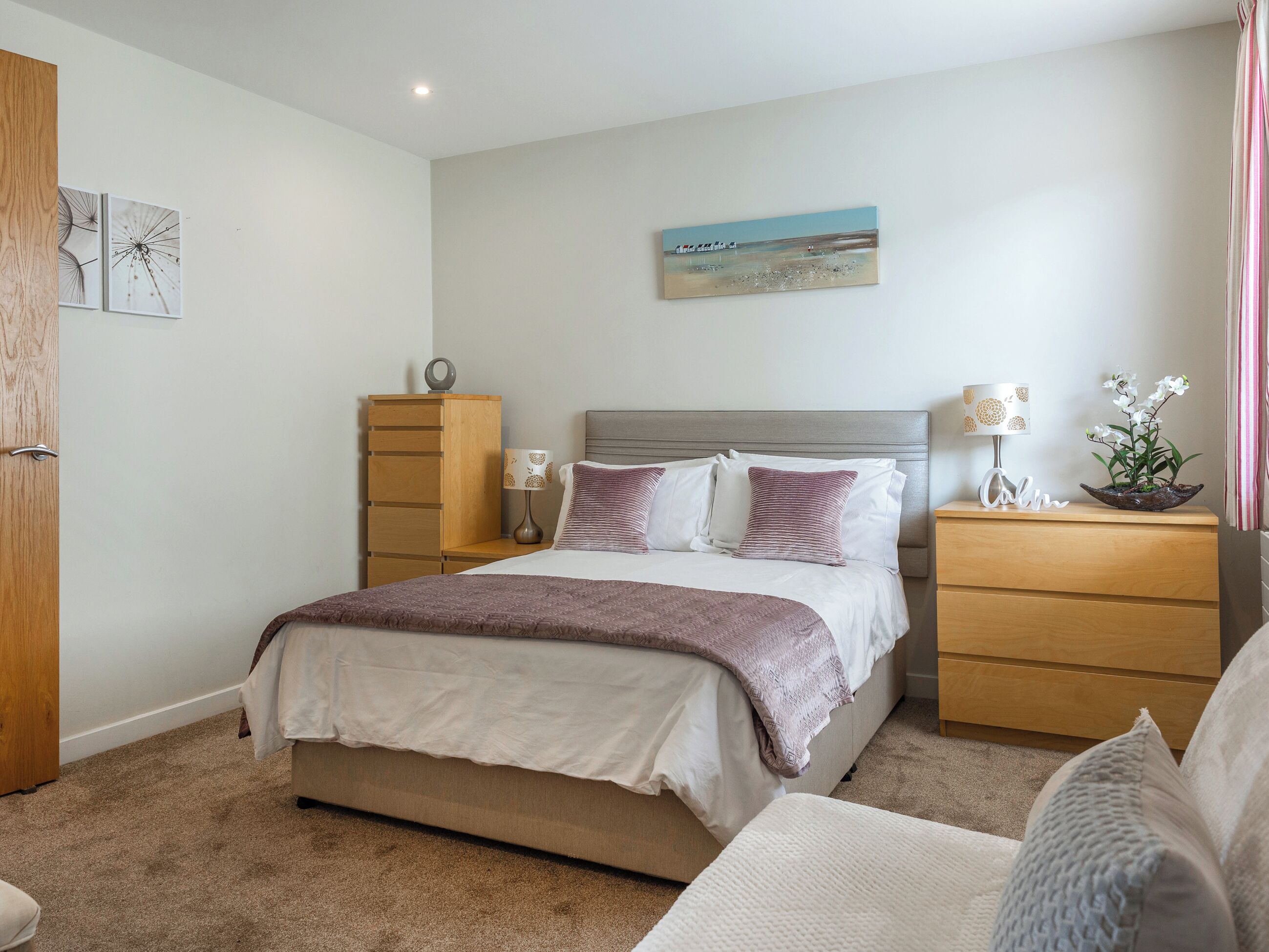 Een slaapkamer met een bed, nachtkastjes, een dressoir, een lamp, een schilderij en een plant.