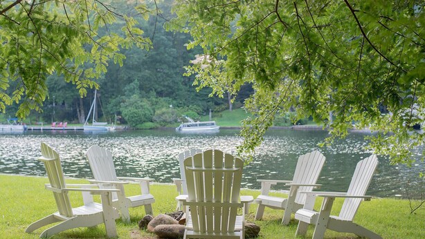 Four Adirondack chairs arranged on a grassy area by a lake with boats in the background.