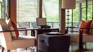 A modern living room with two sofas, a coffee table, and a desk with a lamp. Large windows provide natural light and a view of greenery outside.