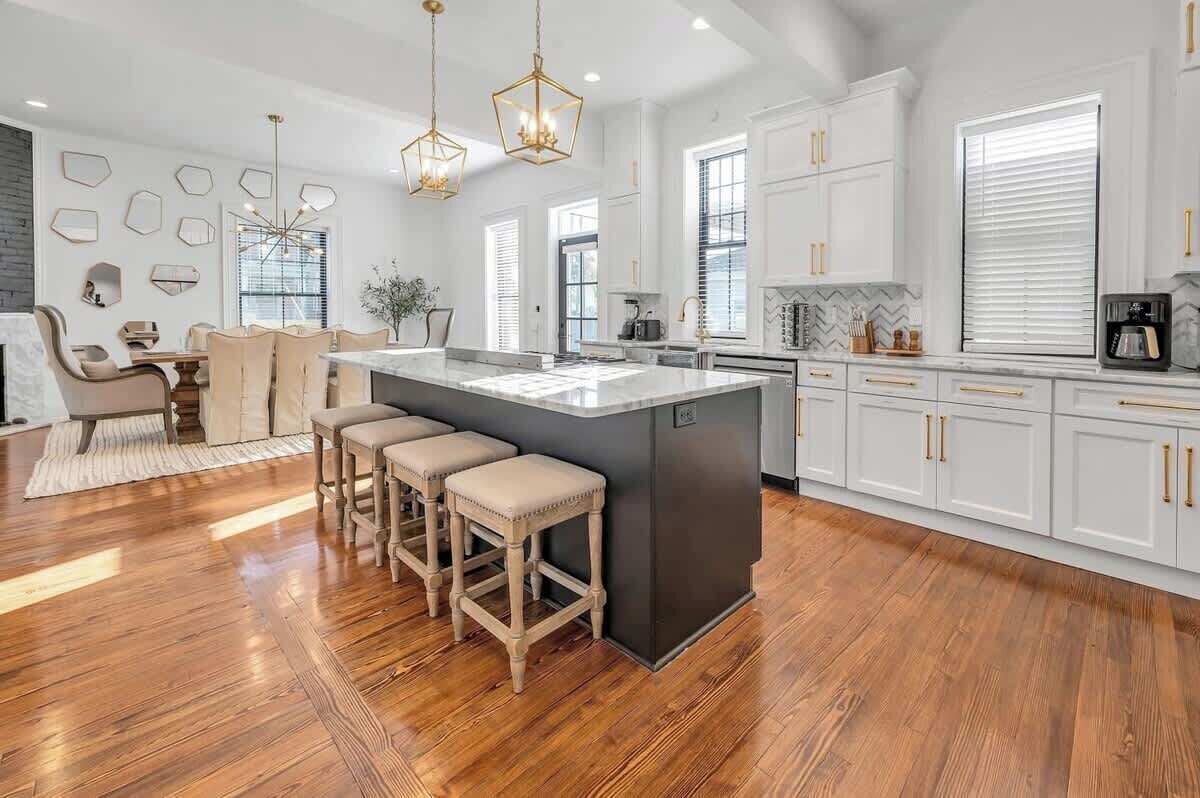Una cocina moderna con armarios blancos, una isla central, taburetes de madera y luminarias colgantes.