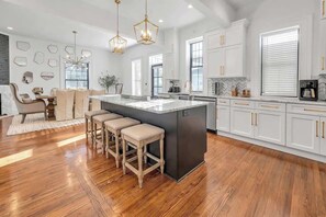 Una cocina moderna con armarios blancos, una isla central, taburetes de madera y luminarias colgantes.