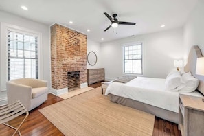 A bedroom with a brick fireplace, a ceiling fan, a bed with white bedding, and a nightstand.