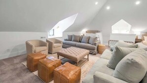 A spacious loft-style room with a high ceiling, a large window, and a cozy seating area with a sofa, armchairs, and ottomans.