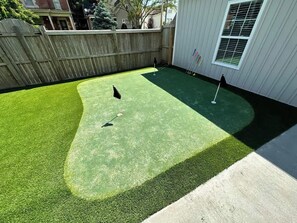 Un jardin avec une pelouse artificielle, une clôture en bois et une maison dotée d’une fenêtre.