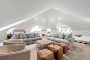Une chambre spacieuse aménagée en loft, avec un plafond haut, une grande fenêtre et un coin salon confortable comprenant un canapé et des poufs.