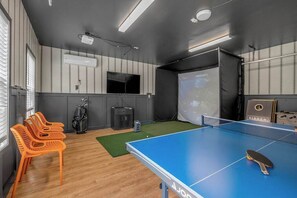 Une table de ping-pong, des chaises, un écran de projection et un haut-parleur dans une pièce dont le sol est en bois et un des murs est gris.