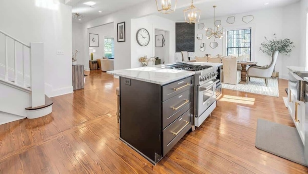 A modern kitchen with a central island, wooden flooring, pendant lighting, and a dining area with a chair and table.