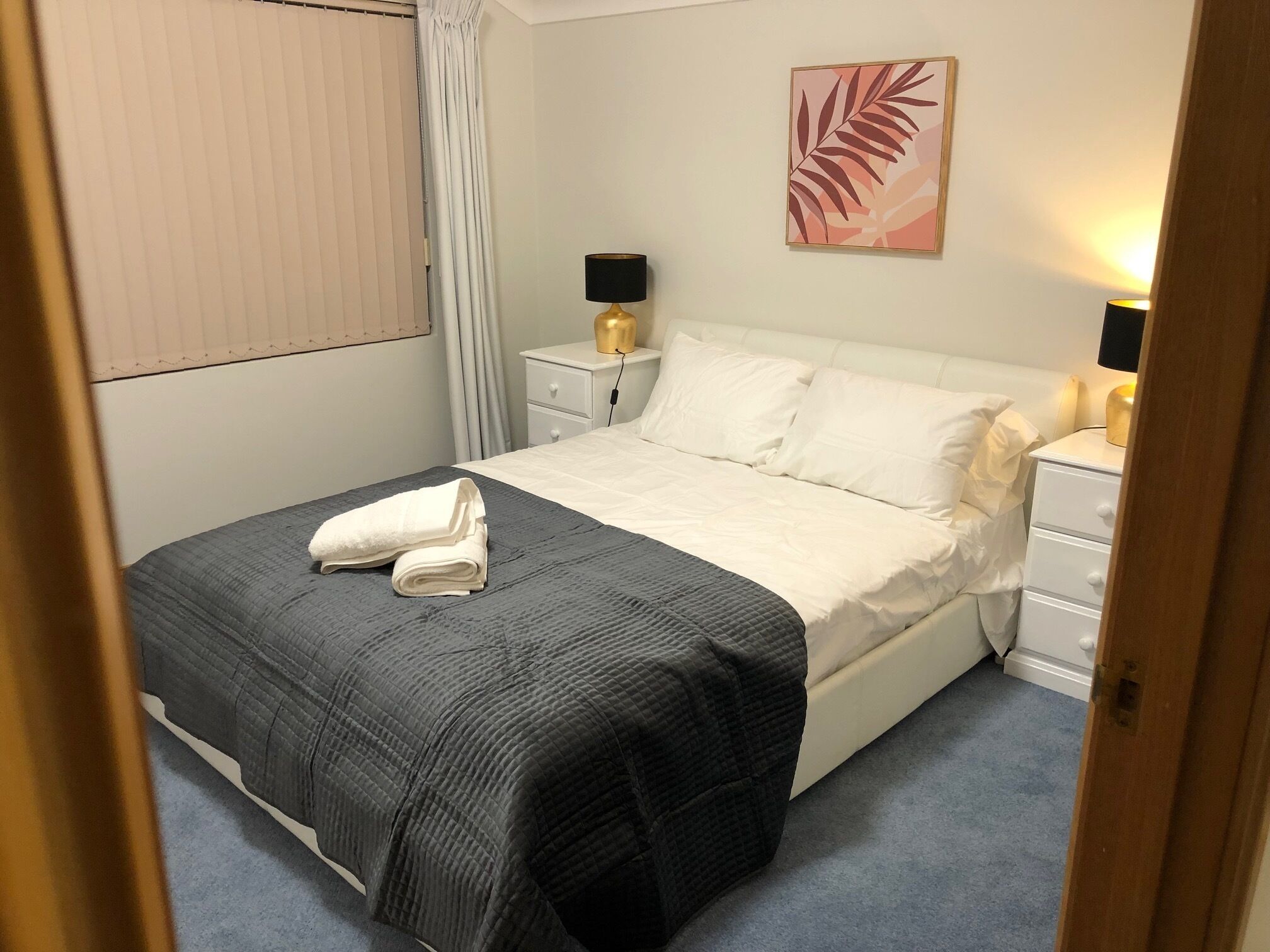 Een netjes opgemaakt bed met een grijze deken, witte kussens en een gevouwen witte handdoek op het voeteneinde. Er is een nachtkastje met een lamp en een kunstwerk van een blad in een lijst aan de muur.
