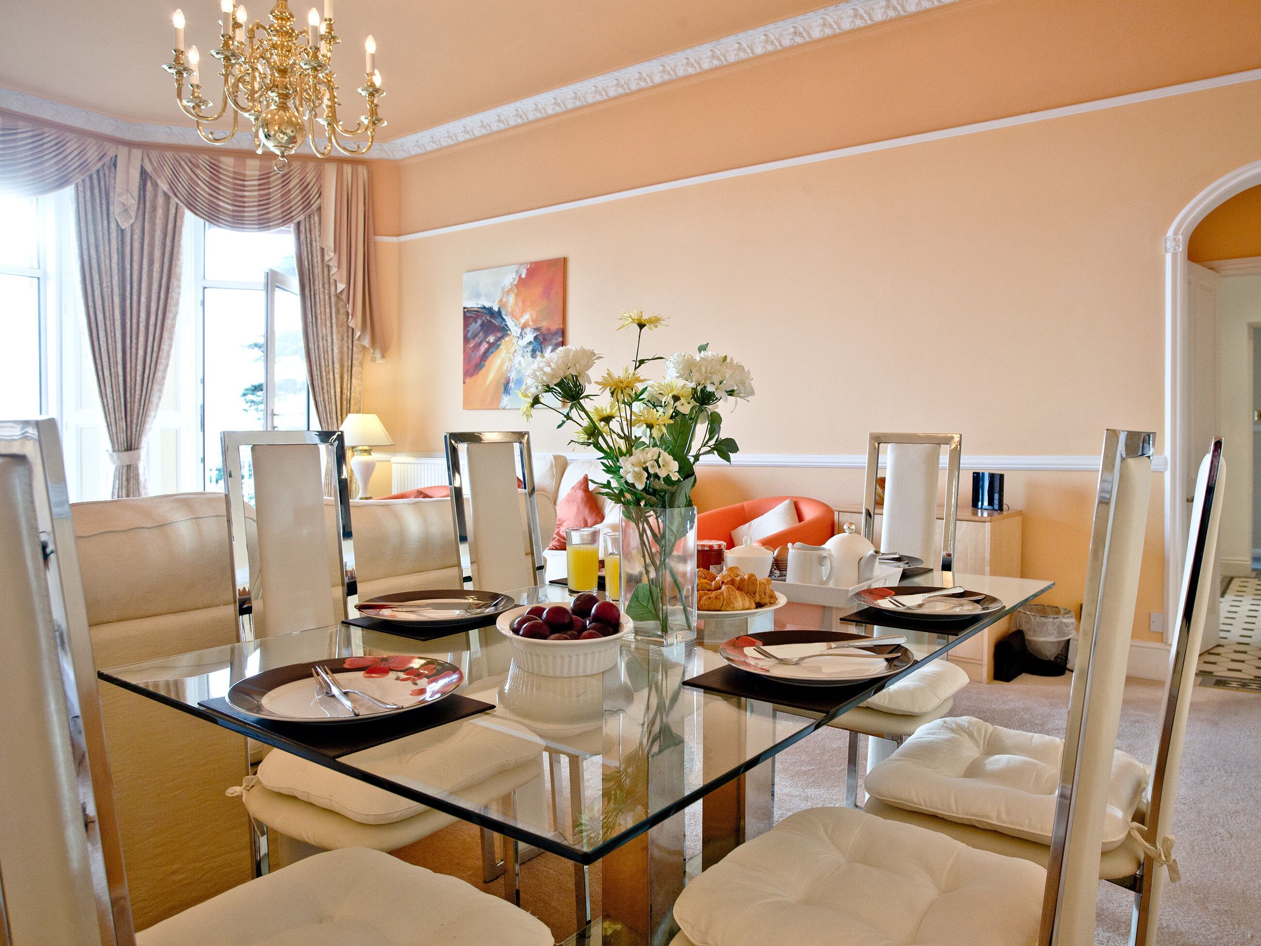 A dining area with a glass table, white chairs, a chandelier, and a vase of flowers.