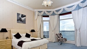 Een ruime slaapkamer met een groot bed, twee nachtkastjes met lampen, een stoel bij het raam en uitzicht op zee.
