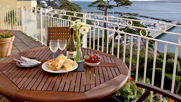 Een houten tafel met een fles wijn, twee glazen, een bord met brood en een schaal fruit op een balkon met uitzicht op een jachthaven en de zee.