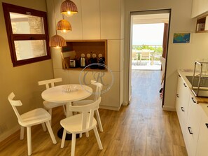 Uma cozinha moderna com armários brancos, uma área de jantar com mesa redonda e cadeiras brancas, e vista para o exterior através de uma porta de vidro.