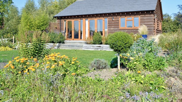 A wooden cabin with a sloped roof, large glass doors, and a well-maintained garden with various flowers and shrubs.
