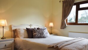A neatly made bed with a white headboard, two bedside tables with lamps, a window with curtains, and a radiator.