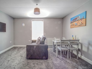Una sala de estar moderna con un sofá gris, una mesa de comedor y sillas. Hay una ventana con persianas, un radiador y un cuadro en la pared.