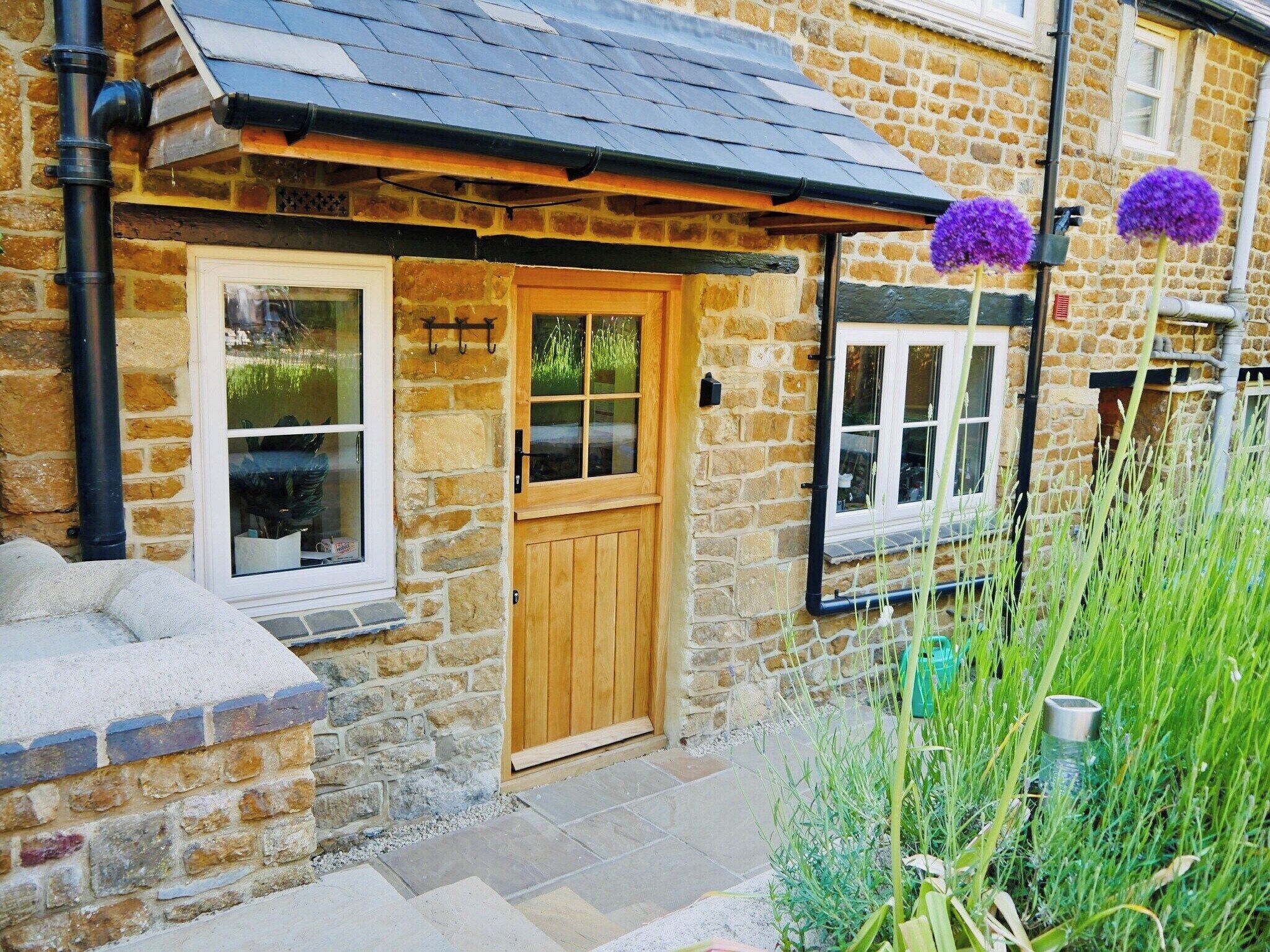 A stone house with a wooden door and windows, a tiled roof, and a garden with plants and a water feature.
