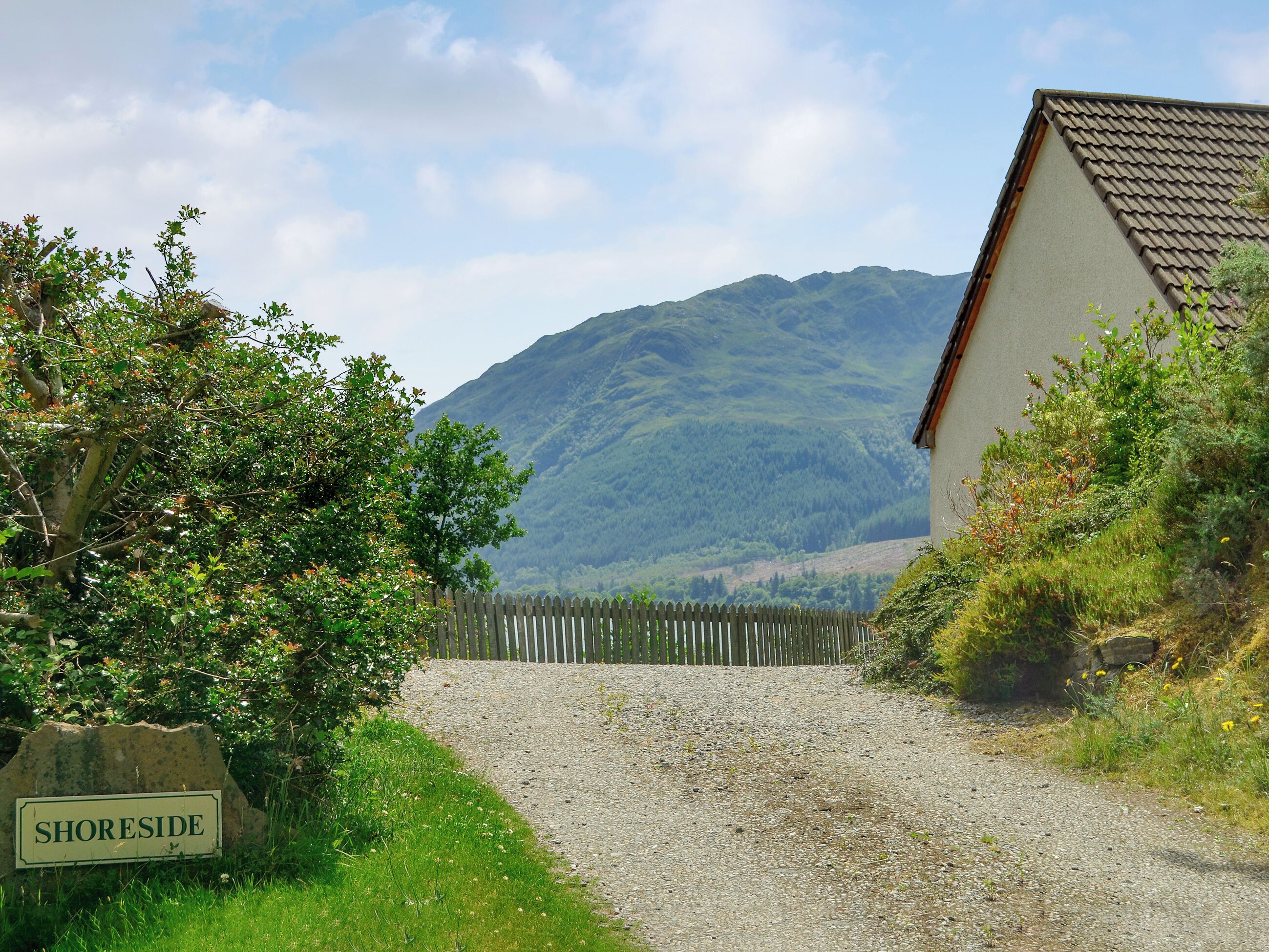 A gravel path leading to a house with a mountain in the background. A wooden fence and a sign reading "SHORESIDE" are visible.
