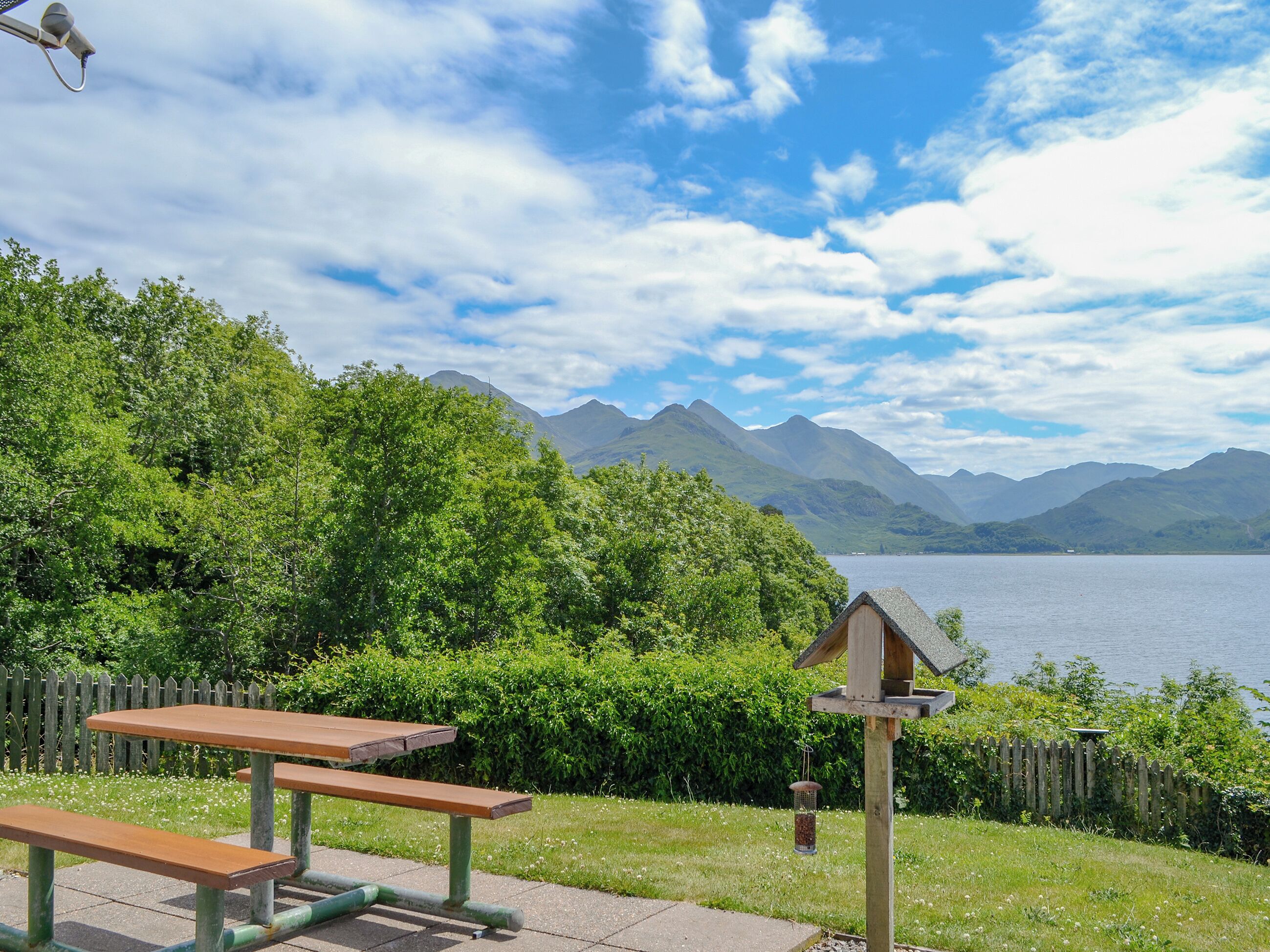 A picnic area with benches and a bird feeder, surrounded by lush greenery and a lake with mountains in the background.