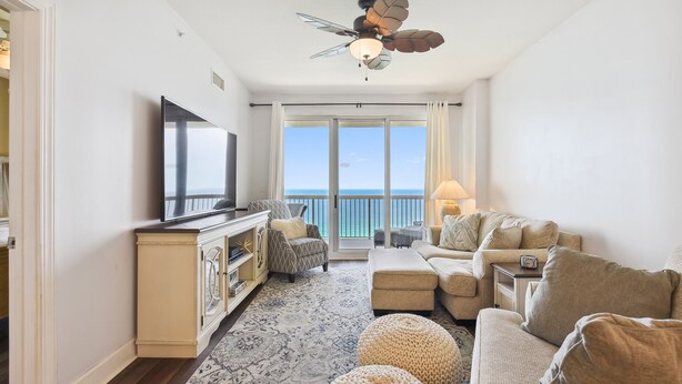 A living room with a flat-screen TV, a ceiling fan, a sofa set, and a view of the ocean through large windows.