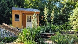 Una pequeña cabaña de madera con techo metálico, rodeada por un jardín con un estanque, rocas y diversas plantas.