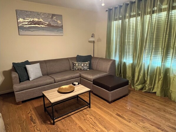 Ein Wohnzimmer mit grauem Sofa, einem kleinen Holztisch und einem grünen Vorhang.