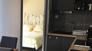 A modern kitchen with dark cabinets, a wooden countertop, and a sink. There is a bed with a floral bedspread in the adjacent room. - APPARTEMENT VACANCES LAGUIOLE (Laguiole)