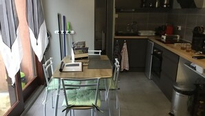 A compact kitchen with a dining area, featuring a table with chairs, a small countertop, and a trash bin. - APPARTEMENT VACANCES LAGUIOLE (Laguiole)