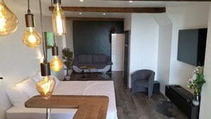 A modern bedroom with a wooden headboard, a bedside table, and a large bed. There is a seating area with a sofa and a small table in the background. - L'essens Fayence... Suite Avec vue Imprenable (Fayence)
