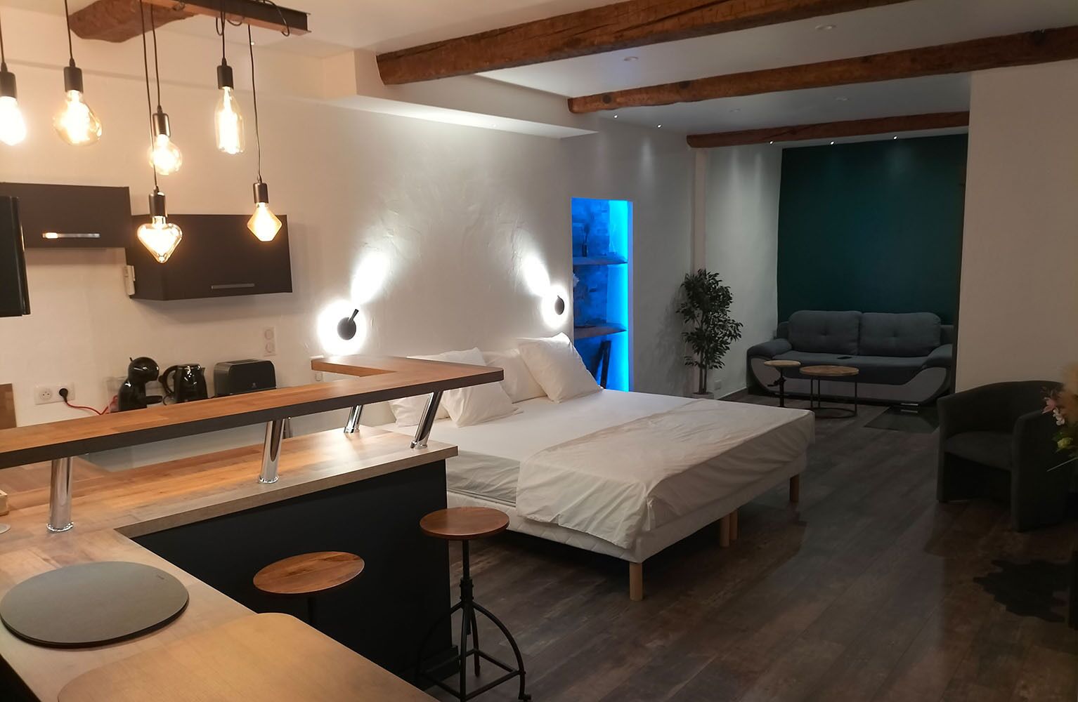 Une chambre d’hôtel moderne comprenant un lit, un petit coin cuisine et un espace salon avec un canapé et des fauteuils.