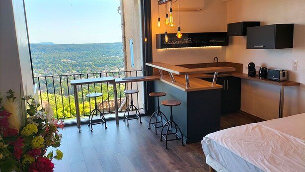 A modern kitchen with a bar area, a large window with a view, and a balcony with a railing and stools. - L'essens Fayence... Suite Avec vue Imprenable (Fayence)