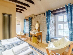 Ein gemütliches Zimmer mit Steinwand, Holzbalken, einem hölzernen Schminktisch mit Spiegel, einem Stuhl und einem Fenster mit blauen Vorhängen.