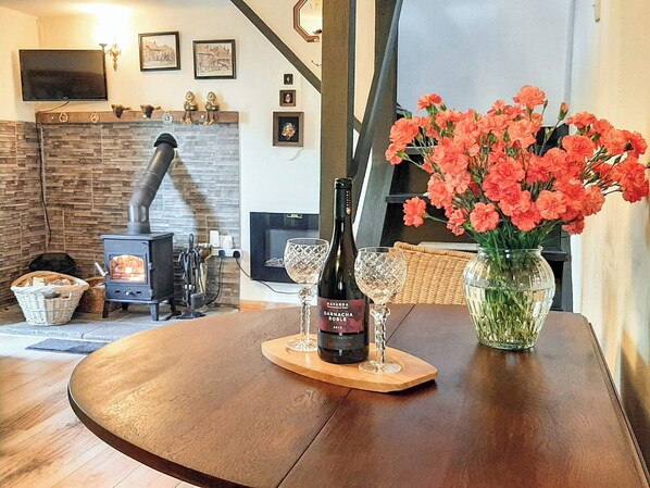 A cozy living room with a stone fireplace, a wooden table with a bottle of wine, a vase of flowers, and a television mounted on the wall.