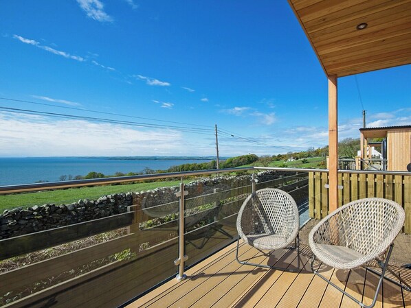 A balcony with two wicker chairs, a wooden deck, and a view of the sea and distant landscape.