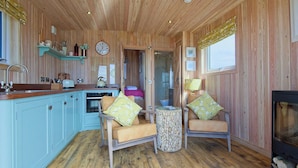 Interior acogedor de una cabaña con techo de madera, una zona de cocina, dos sillas con cojines estampados y una chimenea.